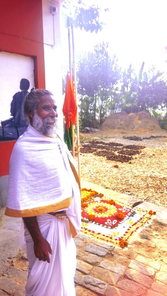 Swamiji Umasankar in white dhoti at Universal Peace Centre ashram, Lachipur during Republic Day 2026 celebrations—smiling beside Sunyoga silhouette mural, saffron flag, marigold rangoli design on sunny grounds with palm trees and cow dung cakes